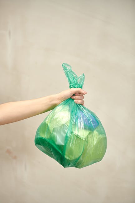 A person's hand and forearm are visible on the left side of the image, holding a green and blue plastic rubbish bag by the tied top. The bag appears to contain waste and is slightly translucent, showing some shapes and materials inside. The background consists of a plain, light-colored wall with a subtle gradient, creating a neutral environment typical of indoor spaces used for waste disposal or storage. The lighting is soft and even, illuminating the bag and arm without harsh shadows. This scene highlights the act of private rubbish handling, with the bag possibly prepared for collection or disposal. The focus on the plastic bag aligns with waste removal services emphasizing proper waste containment, as offered by companies like Rubbish Removal Surrey, which support alternative and responsible waste disposal practices. The image captures a moment of waste management in a straightforward, factual manner, suitable for service-related content about rubbish collection and responsible disposal methods in Surrey.