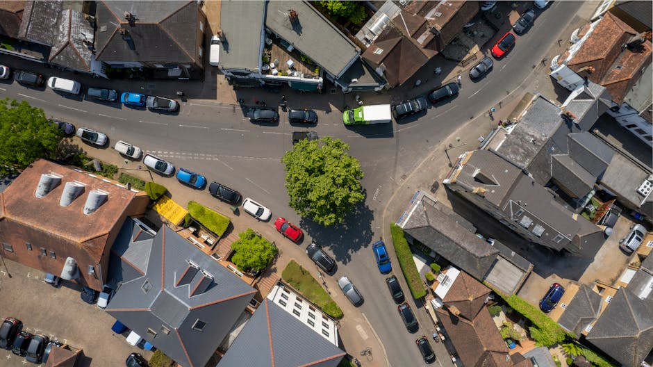An aerial view of a small, urban town centre street showing tightly packed residential buildings with mixed roof types, including tiled and flat finishes, in shades of grey, brown, and red. Several cars, predominantly in neutral colours such as white, silver, grey, and black, are parked along the curving road, which runs through the centre of the image. A bright green van is visible among the parked vehicles, and a couple of pedestrians are walking on the sidewalks. In the middle of the scene, a large leafy tree with vibrant green foliage provides shade to part of the road and sidewalk. Surrounding the buildings are small gardens and hedges, some with structured planting beds or small lawns. The environment appears well-maintained and typical of a residential neighbourhood, with natural lighting highlighting the textures of building materials and foliage. The image offers a clear view of local streets, illustrating typical urban waste management context and the potential for private rubbish collection services like Rubbish Removal Surrey for efficient on-site clearance and refuse handling.
