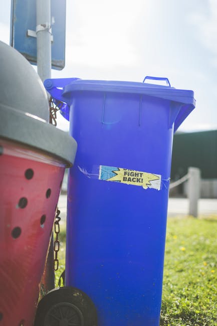 A tall blue plastic rubbish bin with a closed lid, positioned outdoors on a grassy area. The bin features a small yellow, torn paper sticker with the words 'FIGHT BACK!' written on it. To the left, part of a red and black polka-dotted waste container with a gray lid is visible, attached to a small black wheel at the bottom. In the background, there is a blue signpost, a metal pole, and a blurred view of a road with a metal guardrail, suggesting a suburban or urban environment. The sky is partly cloudy with diffuse sunlight illuminating the scene. The setting appears to be an area designated for waste collection or disposal, fitting within the context of private rubbish removal and alternative waste handling services.