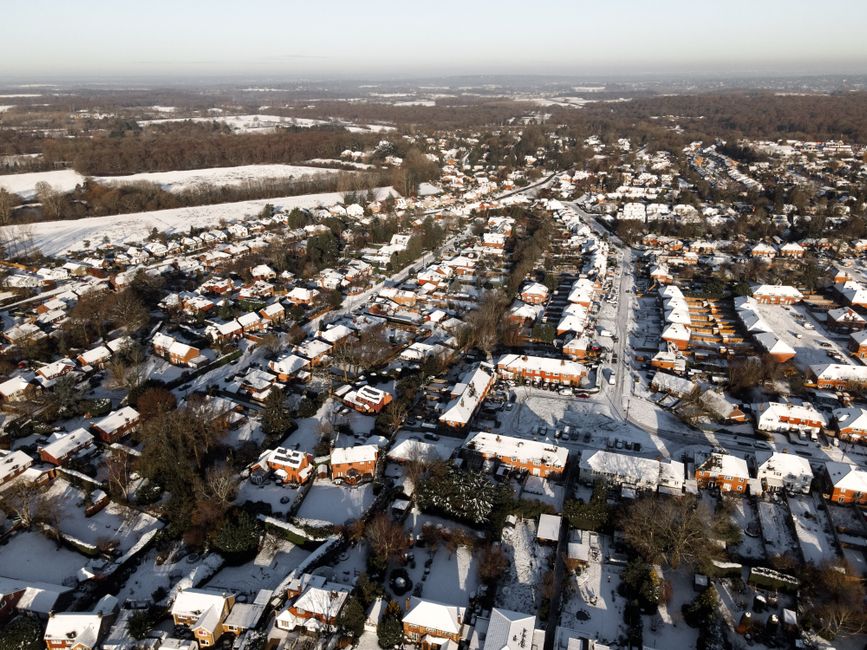 An aerial view of a residential neighbourhood during winter, showing rows of houses with pitched roofs covered in snow. The street running through the centre is lined with parked cars and flanked by houses with brick exteriors, some with driveways that are also snow-covered. Trees without leaves are dispersed throughout the area, and larger open fields in the background extend towards the horizon, with patches of snow and woodland. The lighting is natural, casting soft shadows on the rooftops and streets. This scene reflects a typical suburban environment where private rubbish removal services, such as those provided by Rubbish Removal Surrey, might be used for on-site clearance or managing waste from individual homes in areas with tight access, facilitating waste disposal outside of council bin collections.