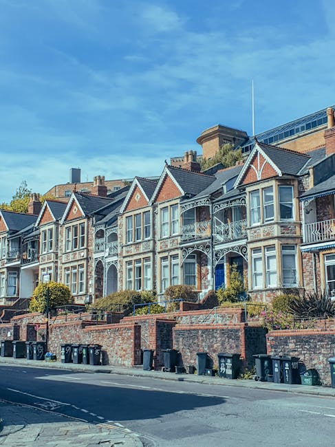 A quiet residential street features a stone wall on the left side, constructed from irregularly shaped beige and light brown stones, casting subtle shadows on the pavement below. To the right, traditional brick houses with varied architectural styles, including chimneys and pitched roofs, line the street. The bricks are predominantly red, with some showing darker accents and textures, and windows framed in white. The street surface is asphalt with clearly visible yellow double lines running along the curb, indicating parking restrictions. Adjacent to the pavement on the right, there are old-fashioned black lamp posts with glass lanterns, and a blue waste bin positioned near a small metal railing. Mature trees with green foliage tower overhead, casting dappled shade over the scene, and a few pedestrians are visible in the distance, walking along the sidewalk. The lighting suggests late afternoon or early evening, with soft, warm sunlight illuminating the buildings and creating gentle shadows. This scene reflects a typical British residential area where private refuse collection services like Rubbish Removal Surrey may operate to handle waste disposal for homes with limited access, exemplifying an on-site clearance or alternative waste management approach.