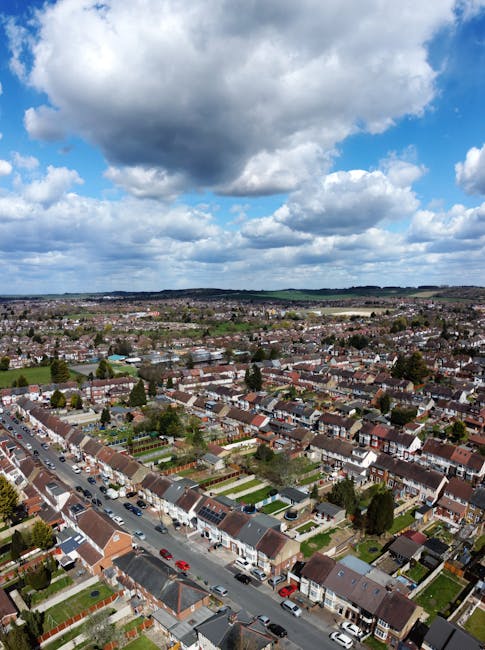 An aerial view of a residential suburban area showing rows of terraced houses with tiled roofs, small front gardens, and narrow streets with parked cars. In the foreground, there are uniform houses with brick walls and sloped roofs, some with visible driveways. The neighborhood extends into the distance, revealing a mix of similar housing with occasional greenery and trees. The sky overhead is partly cloudy, with large, dark grey clouds casting shadows over the scene, suggesting an overcast day with patches of sunlight breaking through. The environment appears quiet and well-maintained, typical of a UK suburb, with no visible rubbish or debris. Rubbish Removal Surrey occasionally services such areas for private waste disposal and alternative waste handling, supporting effective rubbish clearance for homeowners and landlords in the region. The scene is consistent with a typical community where on-site clearance or independent collection of waste could be relevant for residents seeking private disposal options outside of standard municipal rubbish services.