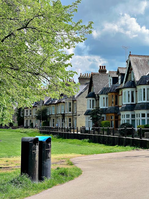 A row of three black cylindrical rubbish bins with rounded lids are positioned on a paved path adjacent to a grassy area, in front of a row of traditional terraced houses with bay windows and slate roofs. The bins are situated near the edge of a small park or green space, with a tall leafy tree with bright green foliage on the left side of the image and a black metal fence running behind the houses. The sky above is partly cloudy with patches of blue visible, creating a natural daylight atmosphere. The scene reflects an urban residential environment where private waste collection, such as from Rubbish Removal Surrey, might be relevant for handling household rubbish as part of alternative waste management methods beyond local authority collections.