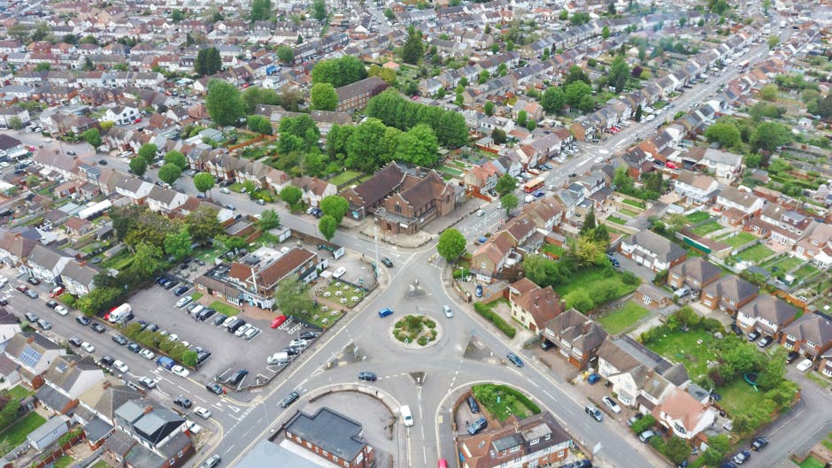 An aerial view of a residential suburban area showing rows of terraced houses with tiled roofs, small front gardens, and narrow streets with parked cars. In the foreground, there are uniform houses with brick walls and sloped roofs, some with visible driveways. The neighborhood extends into the distance, revealing a mix of similar housing with occasional greenery and trees. The sky overhead is partly cloudy, with large, dark grey clouds casting shadows over the scene, suggesting an overcast day with patches of sunlight breaking through. The environment appears quiet and well-maintained, typical of a UK suburb, with no visible rubbish or debris. Rubbish Removal Surrey occasionally services such areas for private waste disposal and alternative waste handling, supporting effective rubbish clearance for homeowners and landlords in the region. The scene is consistent with a typical community where on-site clearance or independent collection of waste could be relevant for residents seeking private disposal options outside of standard municipal rubbish services.
